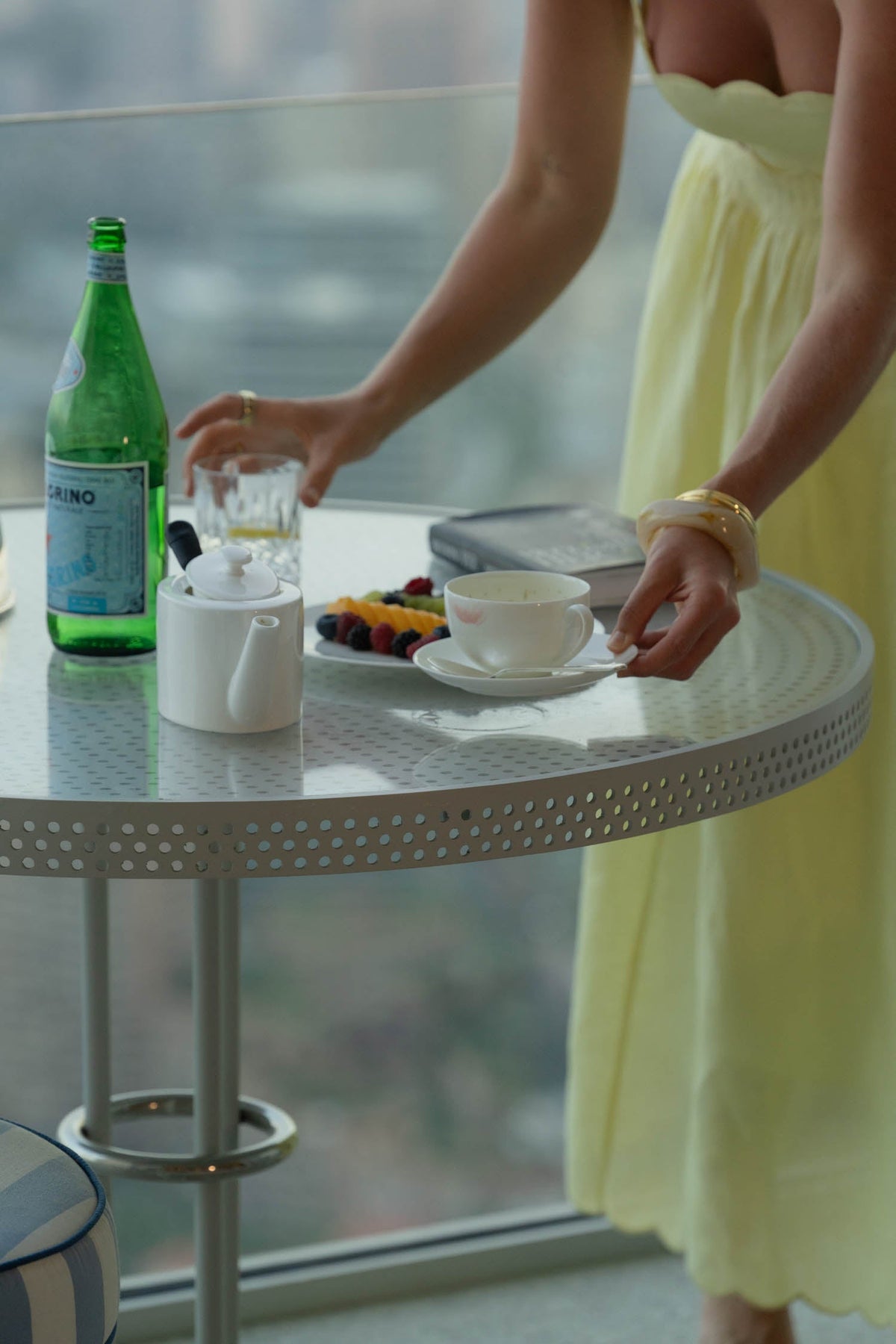 Woman in a yellow dress setting a teacup on a balcony table with fruit, sparkling water and a book